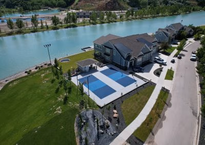 Aerial view of a landscaped residential property in Provo, featuring a large lawn, blue tennis courts, and a river alongside the home, showcasing J&H Landscaping's project work.