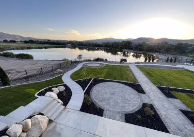 Stunning landscaped yard with stone pathways, circular patio, and lush green grass overlooking a serene lake at sunset, showcasing J&H Landscaping's expertise in Provo, UT.