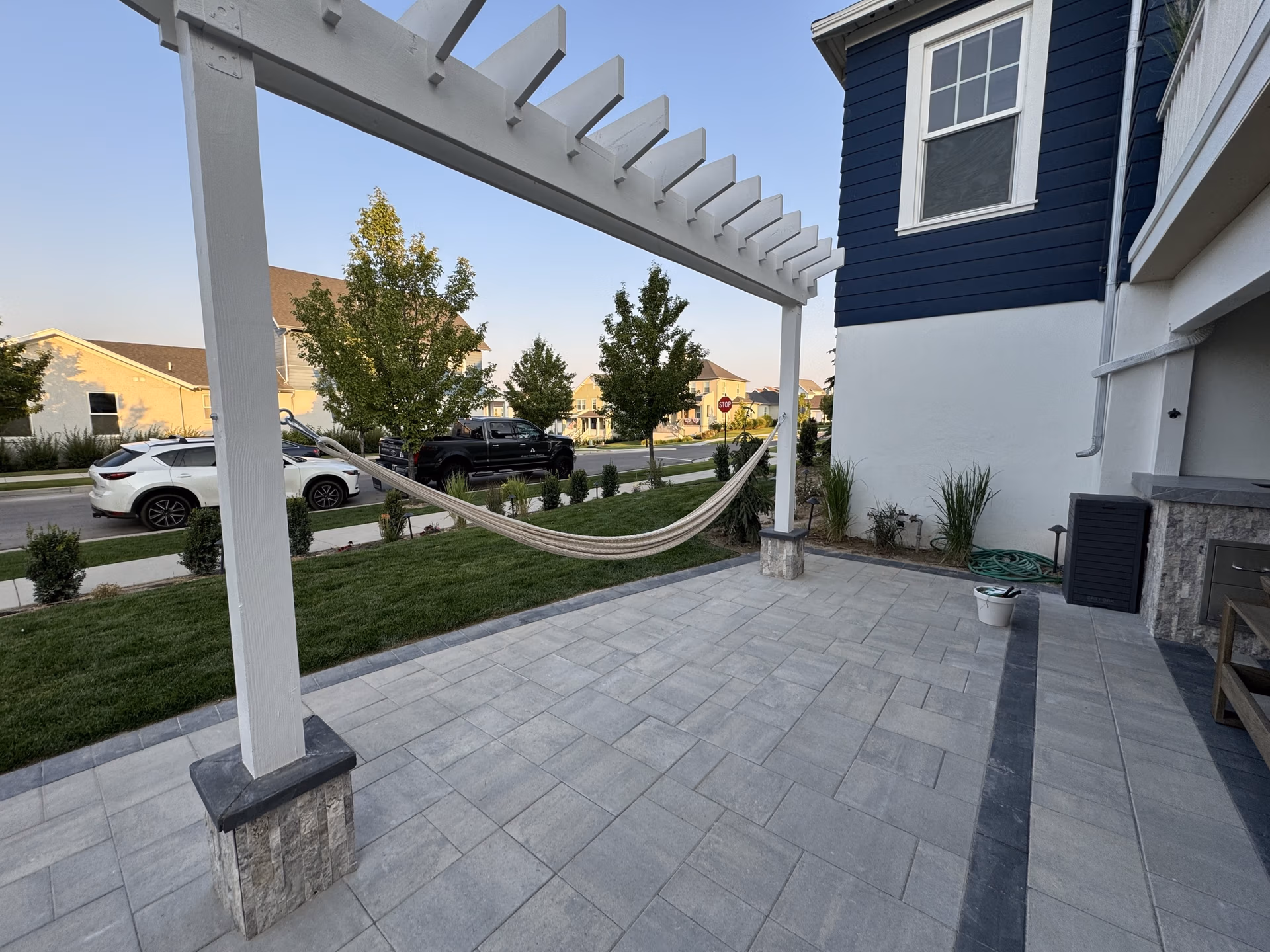 Patio with stone pavers, white pergola, and hammock, surrounded by landscaped greenery, showcasing durable outdoor space designed for relaxation in Spanish Fork, Utah.