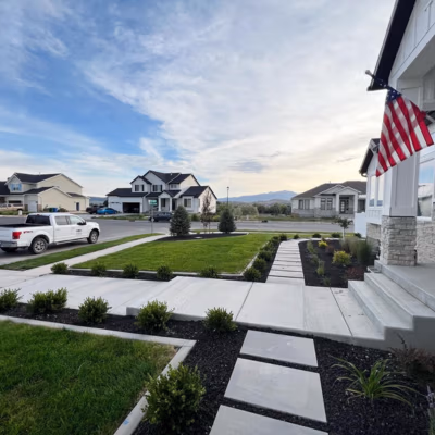 Lawn and landscaped front yard in Lindon, Utah, featuring clean concrete pathways, trimmed bushes, and a flag, showcasing J&H Landscaping's quality and attention to detail.