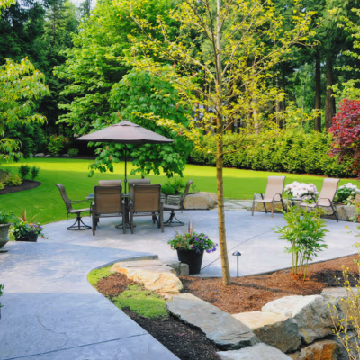 Lush, well-maintained backyard with green lawn, patio area featuring a table and chairs under an umbrella, surrounded by trees and flowering plants, ideal for Utah homeowners seeking reliable landscaping services.