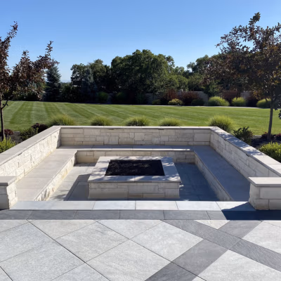Modern concrete fire pit surrounded by a stone seating area, set in a landscaped yard with green grass and trees, showcasing durable hardscaping design for outdoor spaces in Mapleton.